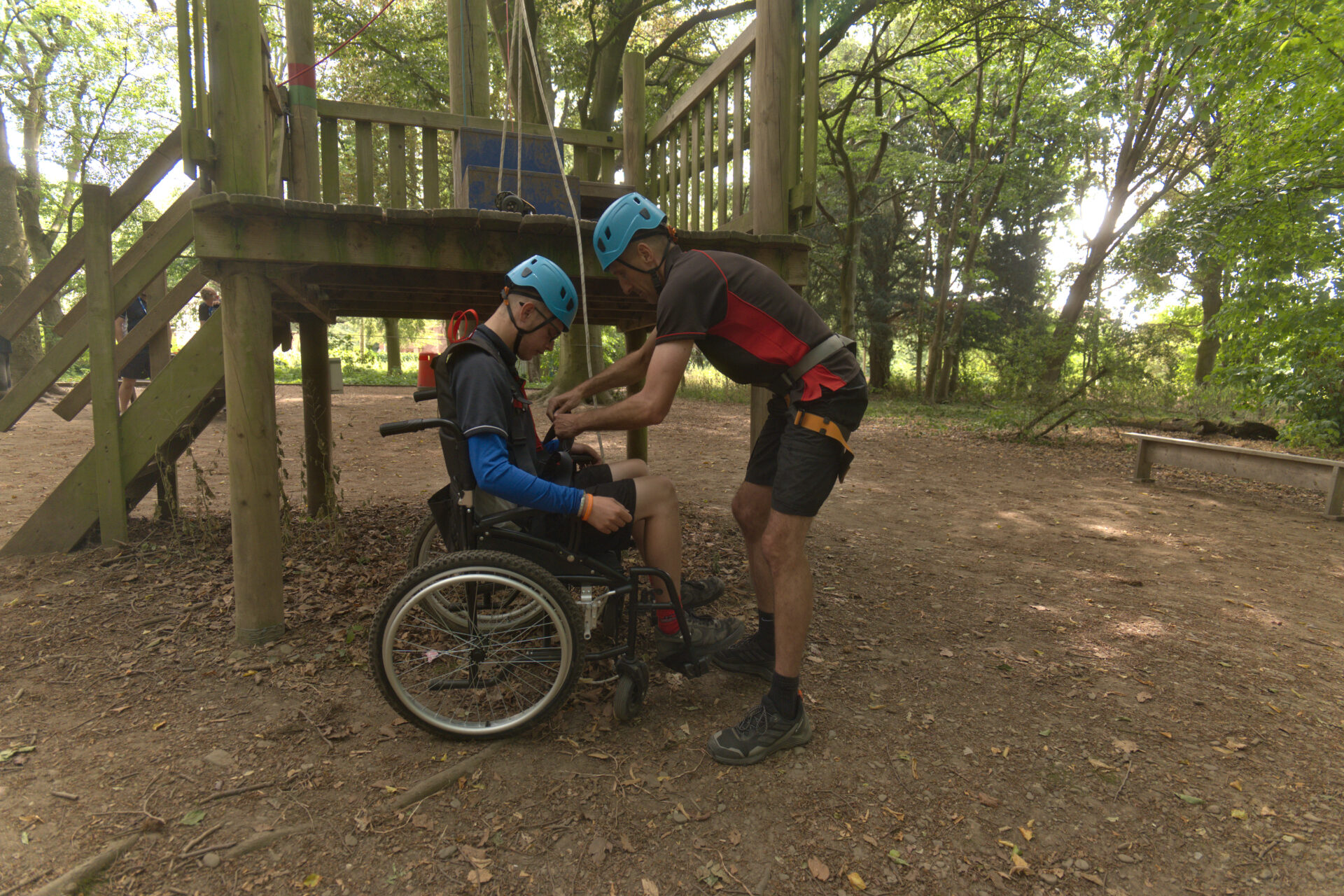 Instructor fitting a harness for a wheelchair user beneath a high ropes platform in the woods
