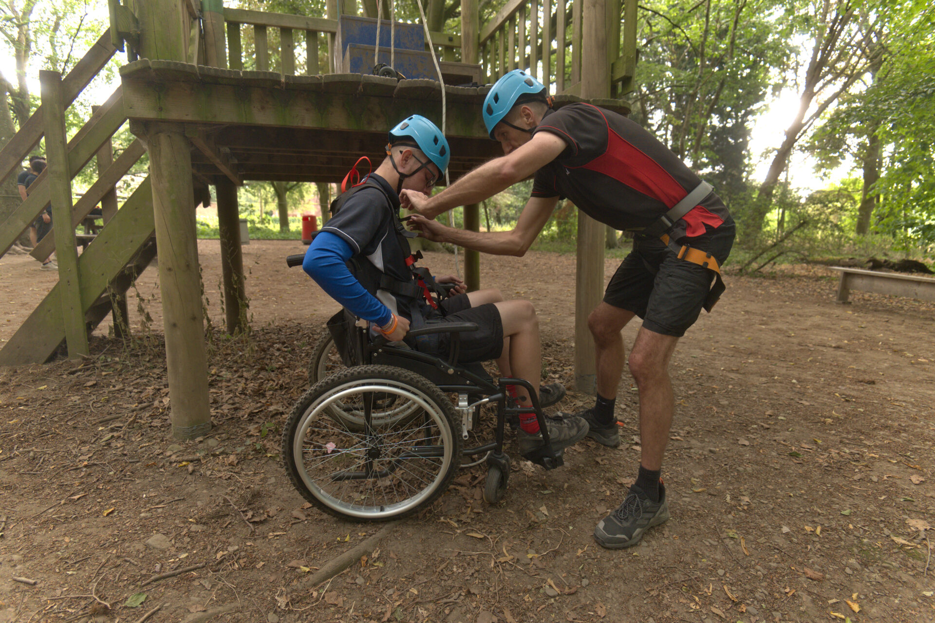 Instructor fastening an adapted harness for a young wheelchair user under a woodland platform