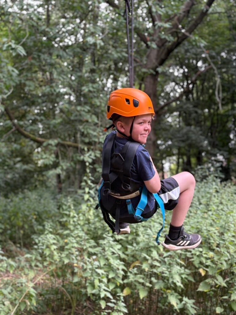 A young boy in an orange helmet, smiling, on a zip line.